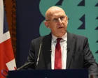 John Healey hablando en un atril con banderas del Reino Unido al fondo